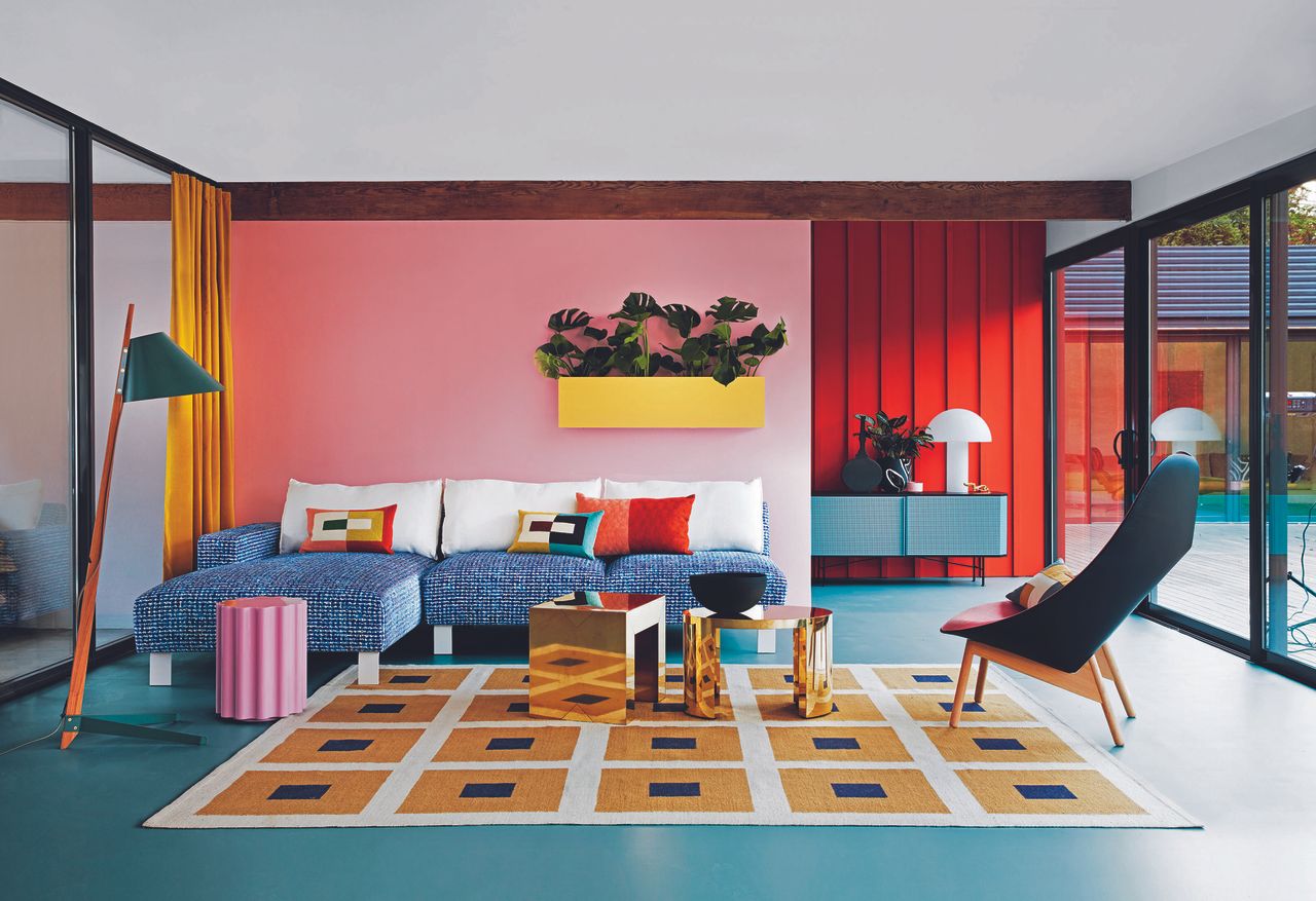 Living room with colourful L shaped sofa and cushions, walls in red and pink with glass doors either side and pink and gold occasional tables