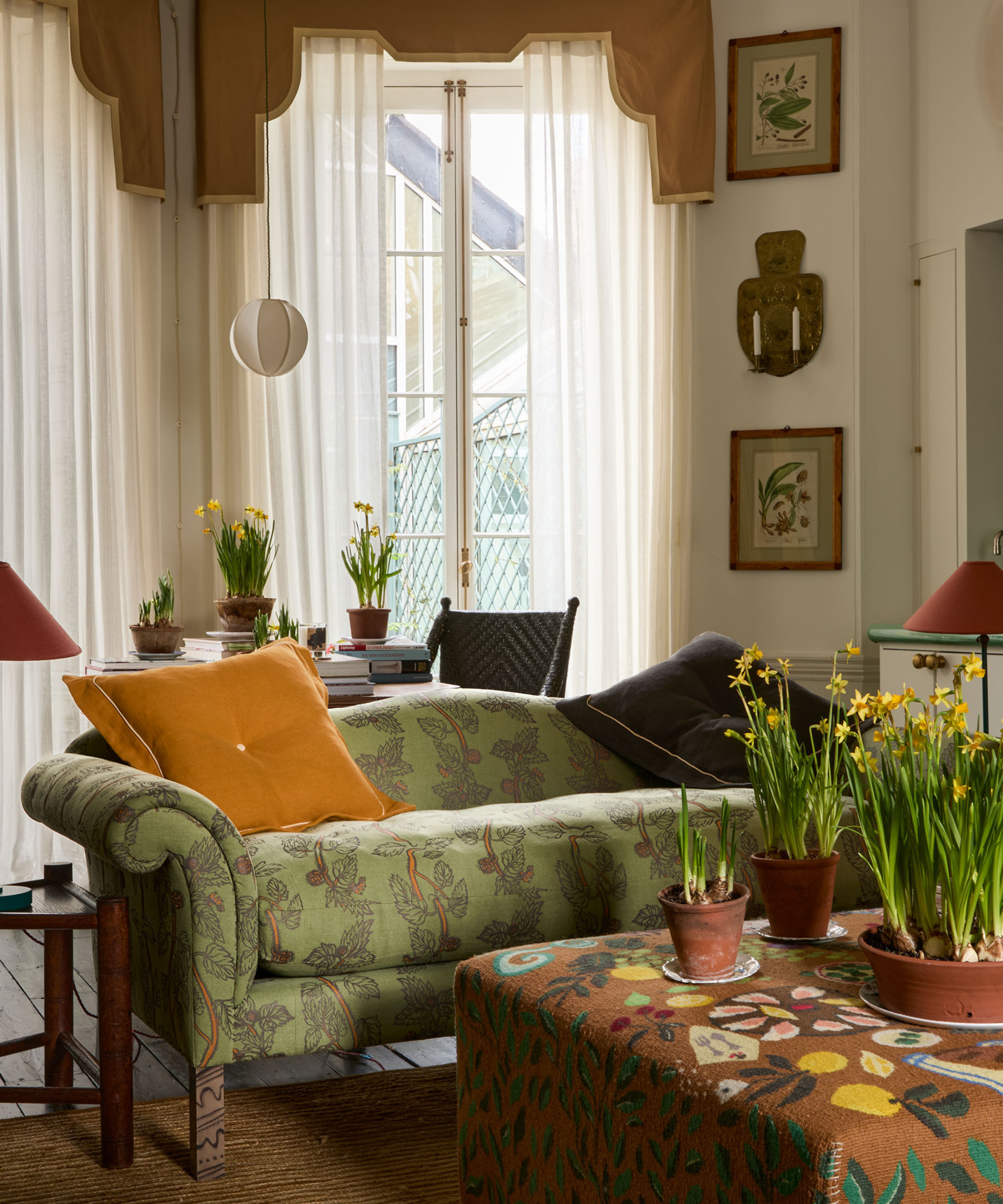 a cozy living room with a green printed sofa, a floral printed ottoman, plants, and sheer drapes in the background