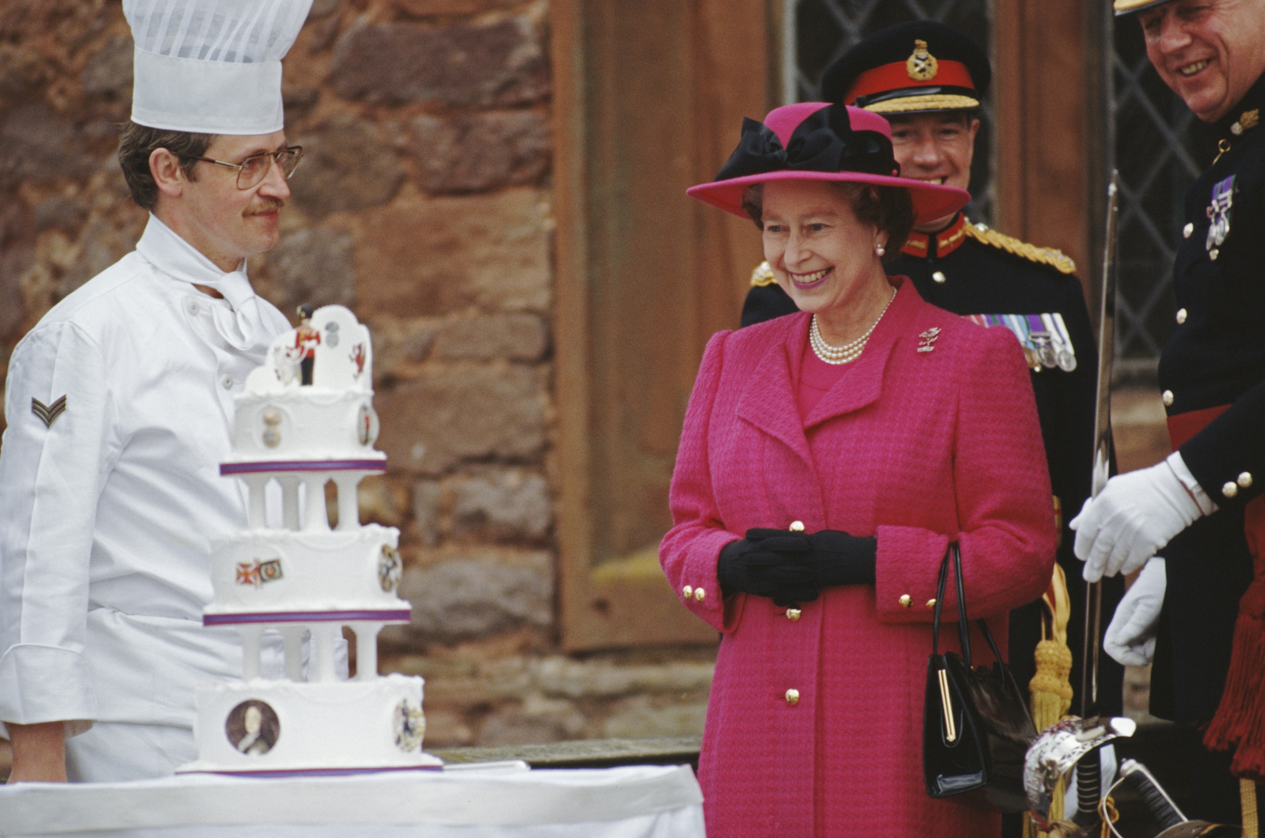 Queen Elizabeth wearing a pink coat smiling at her birthday cake with a chef standing to the side