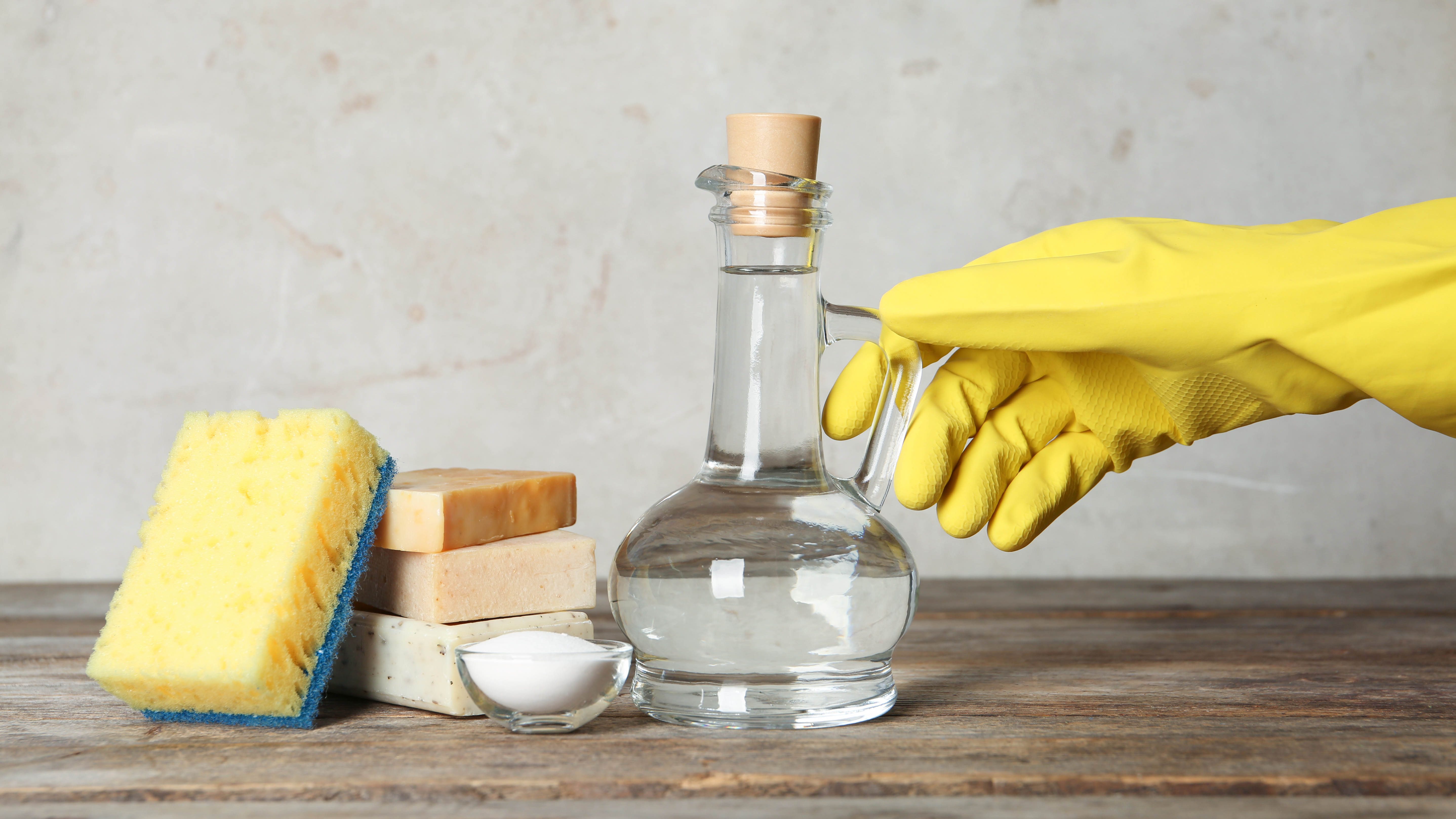 A gloved hand reaches for a stoppered jug of distilled white vinegar next to a sponge and baking soda