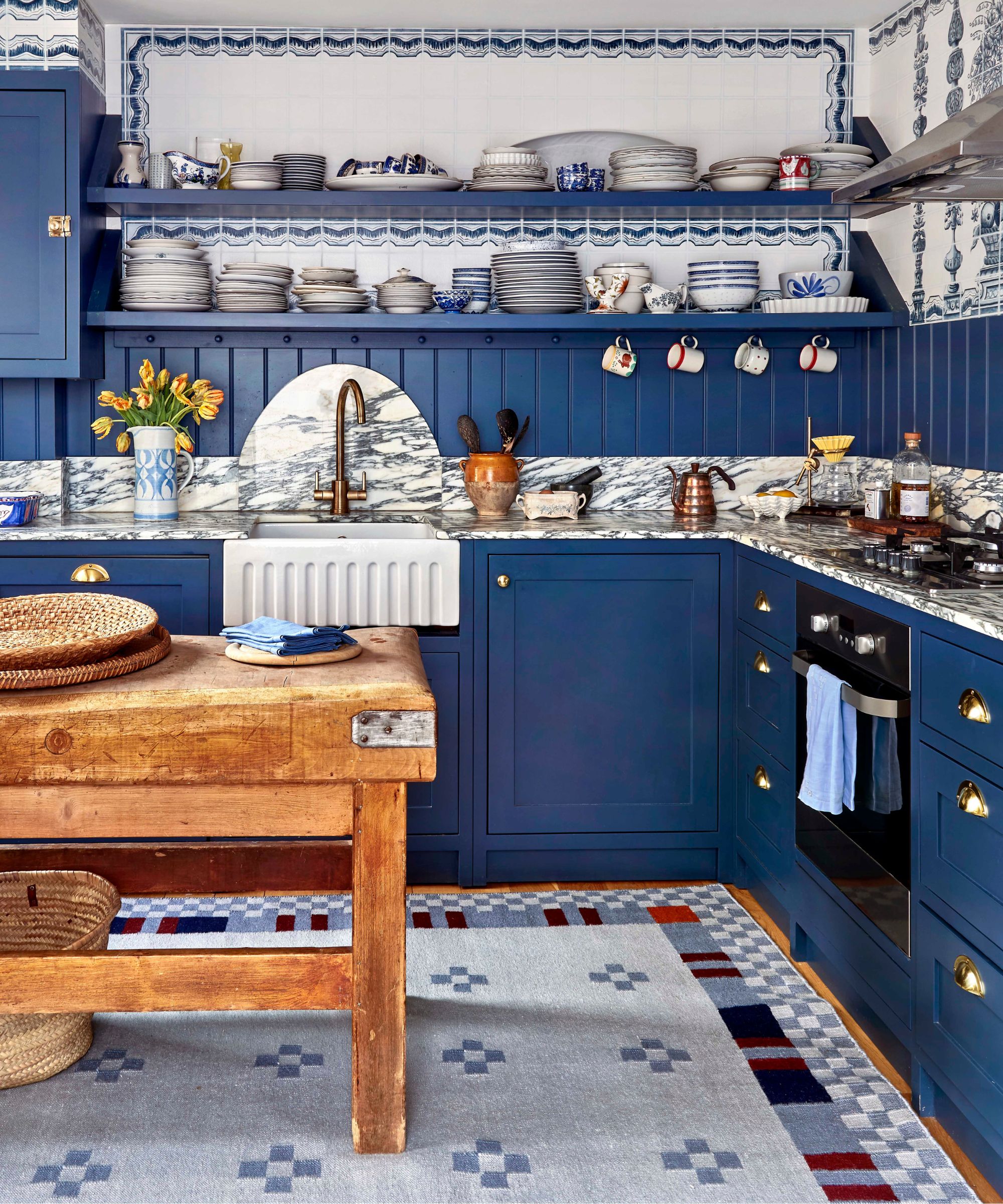 blue scandinavian maximalist kitchen styled with marble countertops, a fluted sink, open shelving and scalloped tiles with a wooden butcher block island stood on a folky blue patterned rug
