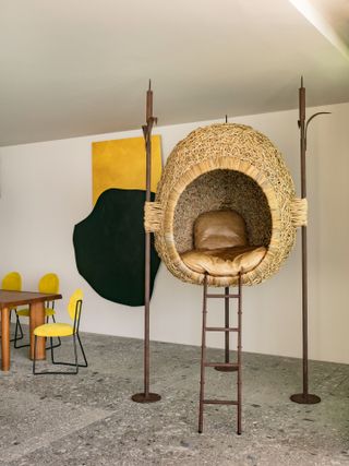 a wooden nest sculpture with a ladder leading up to it with cushions inside, in a room with grey marble flooring and a dining table with mustard yellow chairs in the background