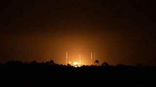 a rocket launches at night in the distance, silhouetting trees in between it and the photographer