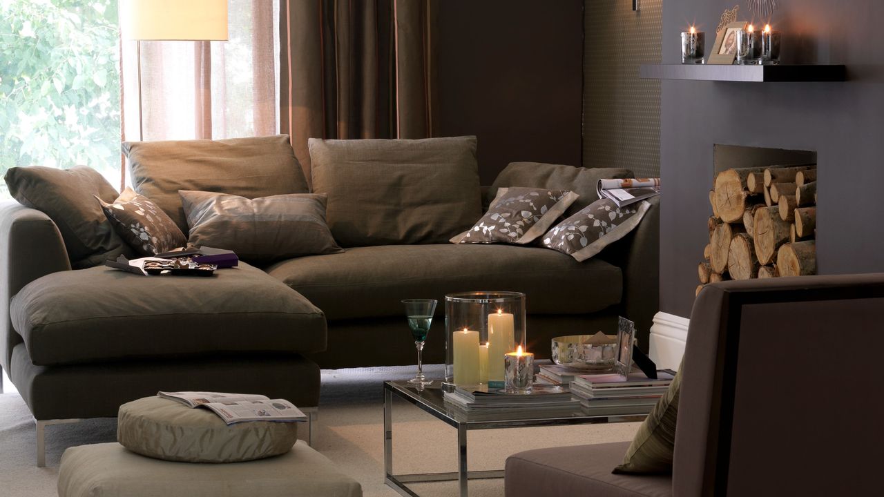 A brown-painted living room with a brown chaise sofa and candles lit on a glass coffee table