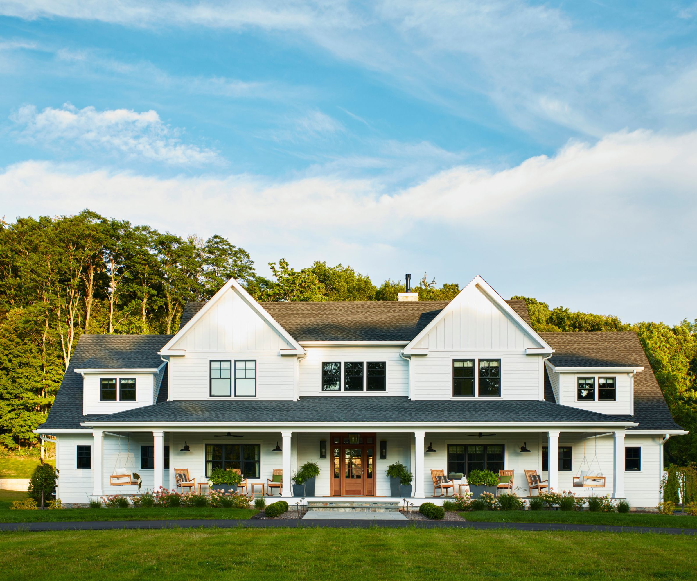 exterior of a white farmhouse building designed by camden grace interiors with a large porch styled with rocking chairs, swing porch chairs and traditional planting