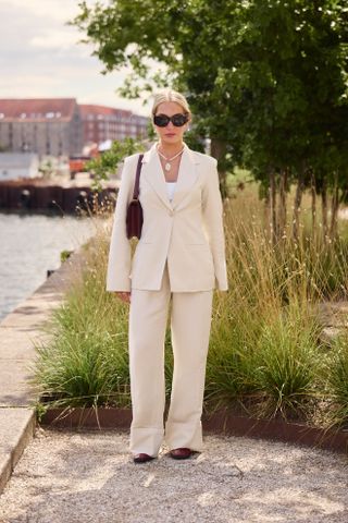 A guest attends the Copenhagen spring summer fashion week wearing a white suit set, black sunglasses, and shoulder bag