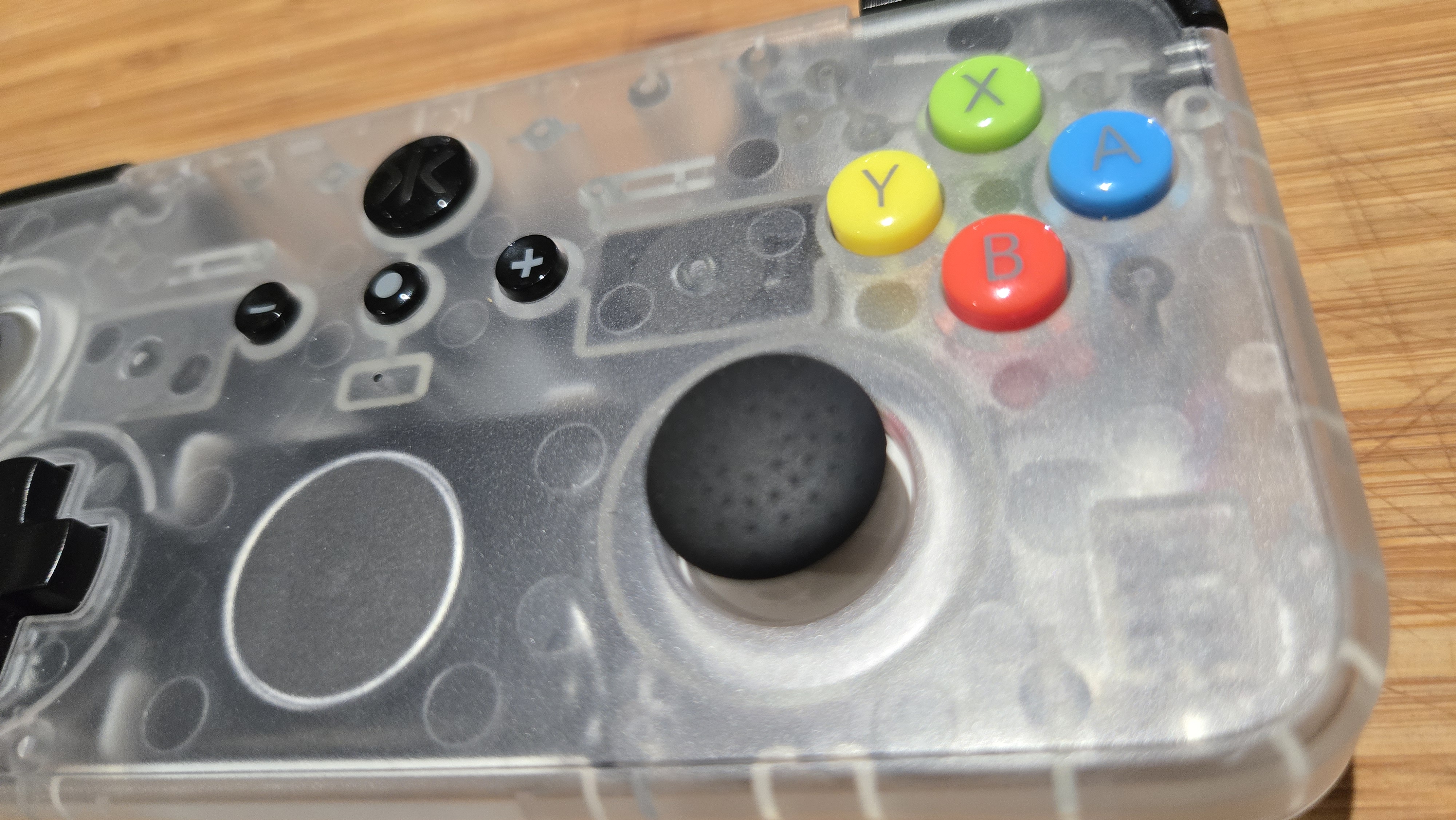 A close up of a Classic Clear edition of the CRKD NEO S rectangular controller. The frame focusses primarily on the right thumbstick and the face buttons. Alongside other fixtures and fittings in black, the colourful face buttons stand out: the leftmost 'Y' button is yellow, the topmost 'X' button is green, the rightmost 'A' button is blue, and the bottommost 'B' button is red. This design flourish is also notable as the main body of the controller is made out of a grey-ish cloudy clear plastic that allows the viewer to see the controller's internals.