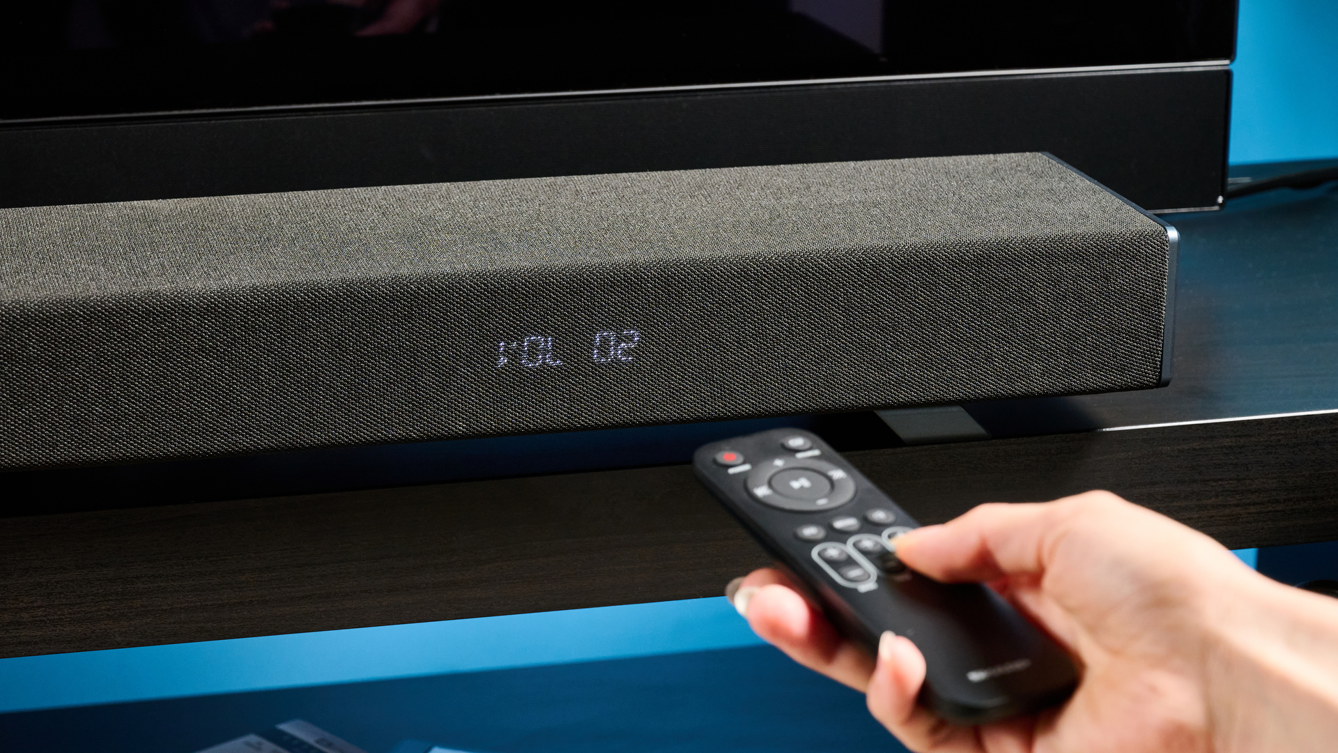 the Sharp 3.1.2 HT-SBW53121 Soundbar in black with a remote, subwoofer, against a blue background next to a panasonic TV