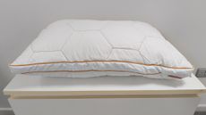 A white bed pillow with a geometric hexagonal quilted pattern and orange piping sitting on a white surface.