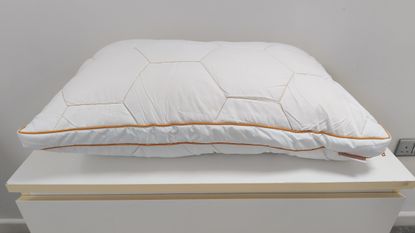 A white bed pillow with a geometric hexagonal quilted pattern and orange piping sitting on a white surface.