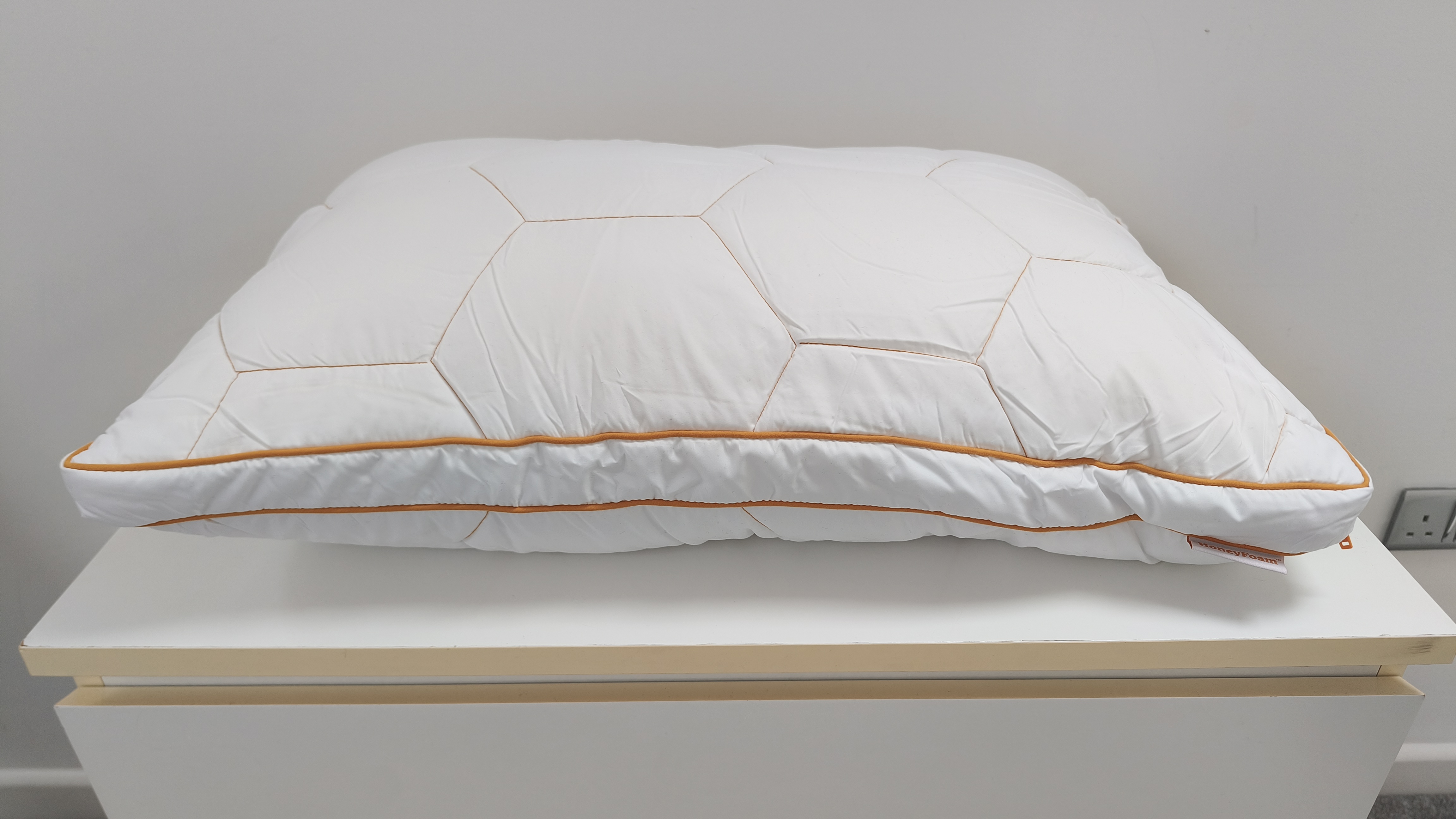 A white bed pillow with a geometric hexagonal quilted pattern and orange piping sitting on a white surface.