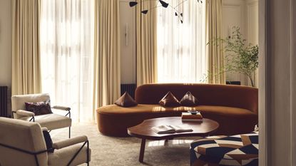 Image of a burnt orange, curved sofa next to two white boucle accent chairs and a wooden coffee table in a white living room with high ceilings and two large windows with off-white curtains.