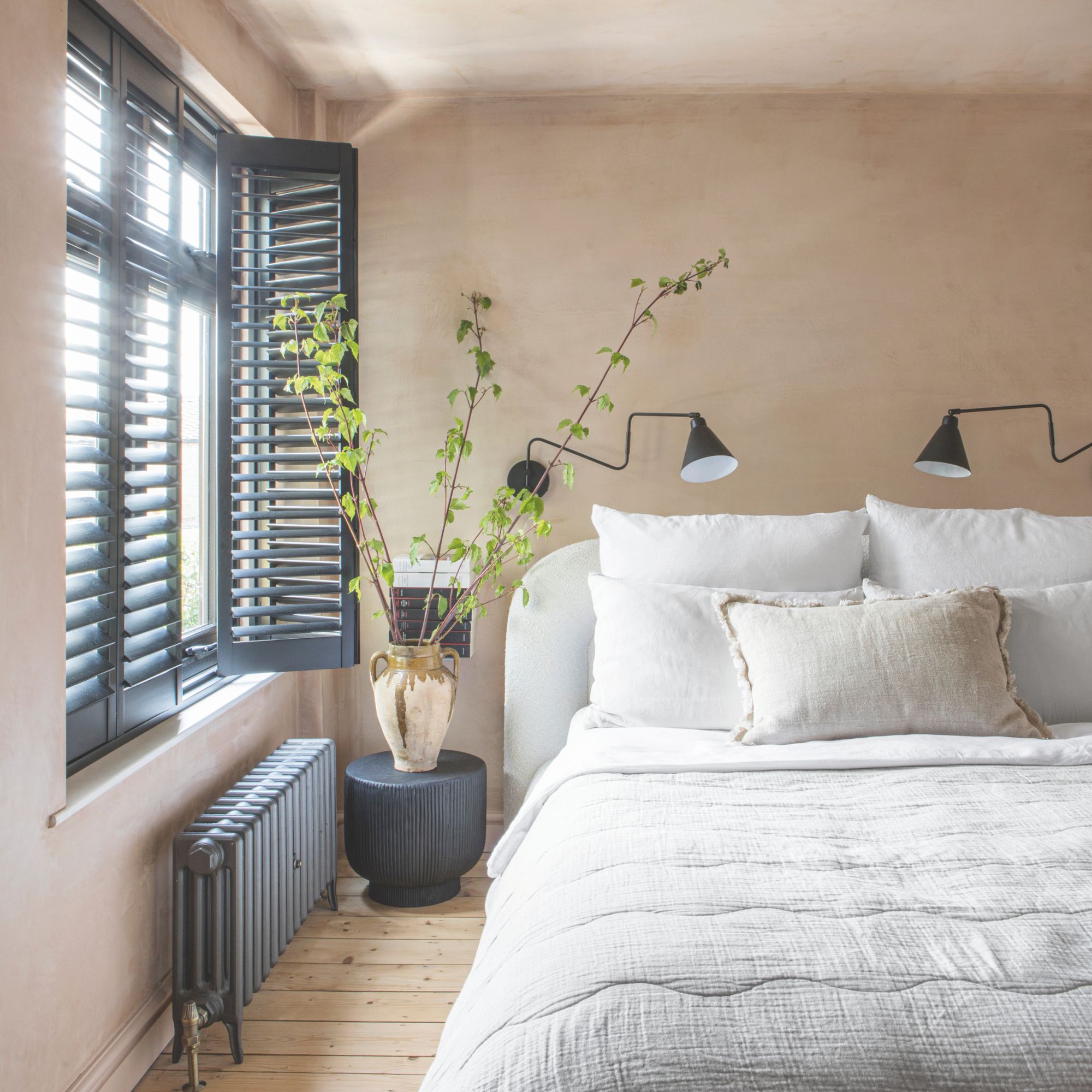 Bedroom painted in plaster-pink paint with a large bed with white bedding on it, and black shutters on the window