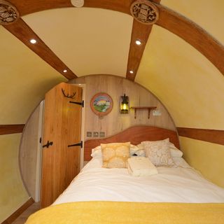 hobbit house bedroom showing the bed with white and yellow coverings up against a wooden wall with a wooden door, and curved yellow walls with wooden beams