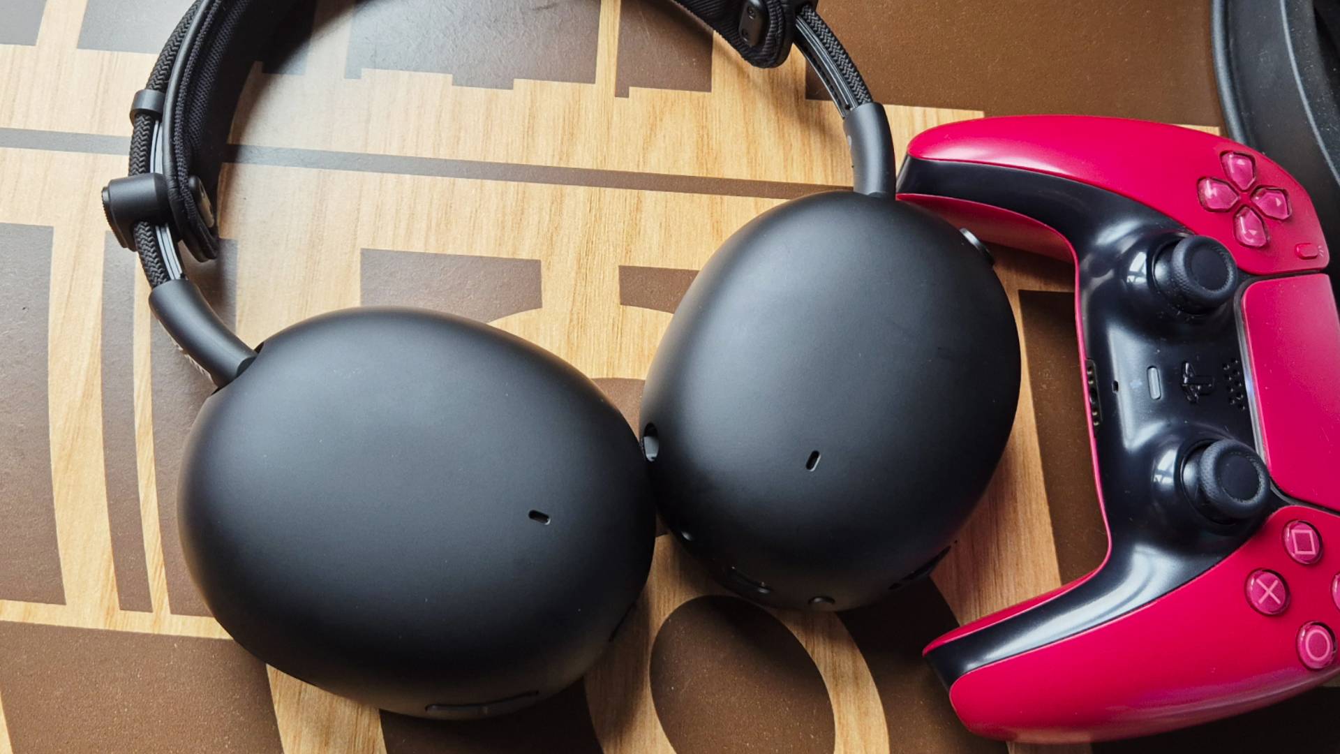 Photo of a black headset sitting on a table with a red PS5 DualSense controller to the right of the frame.