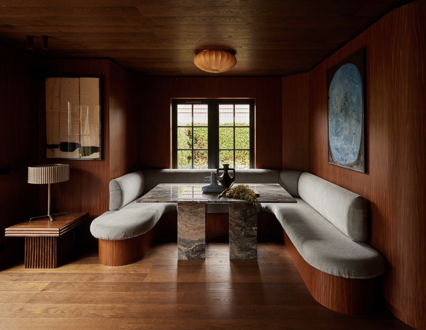 Image of a breakfast nook drenched in wood paneling, wood flooring, and wooden ceilings. There is a light gray bench seat with a wooden base that goes around the entire nook and a square marble table in the center.