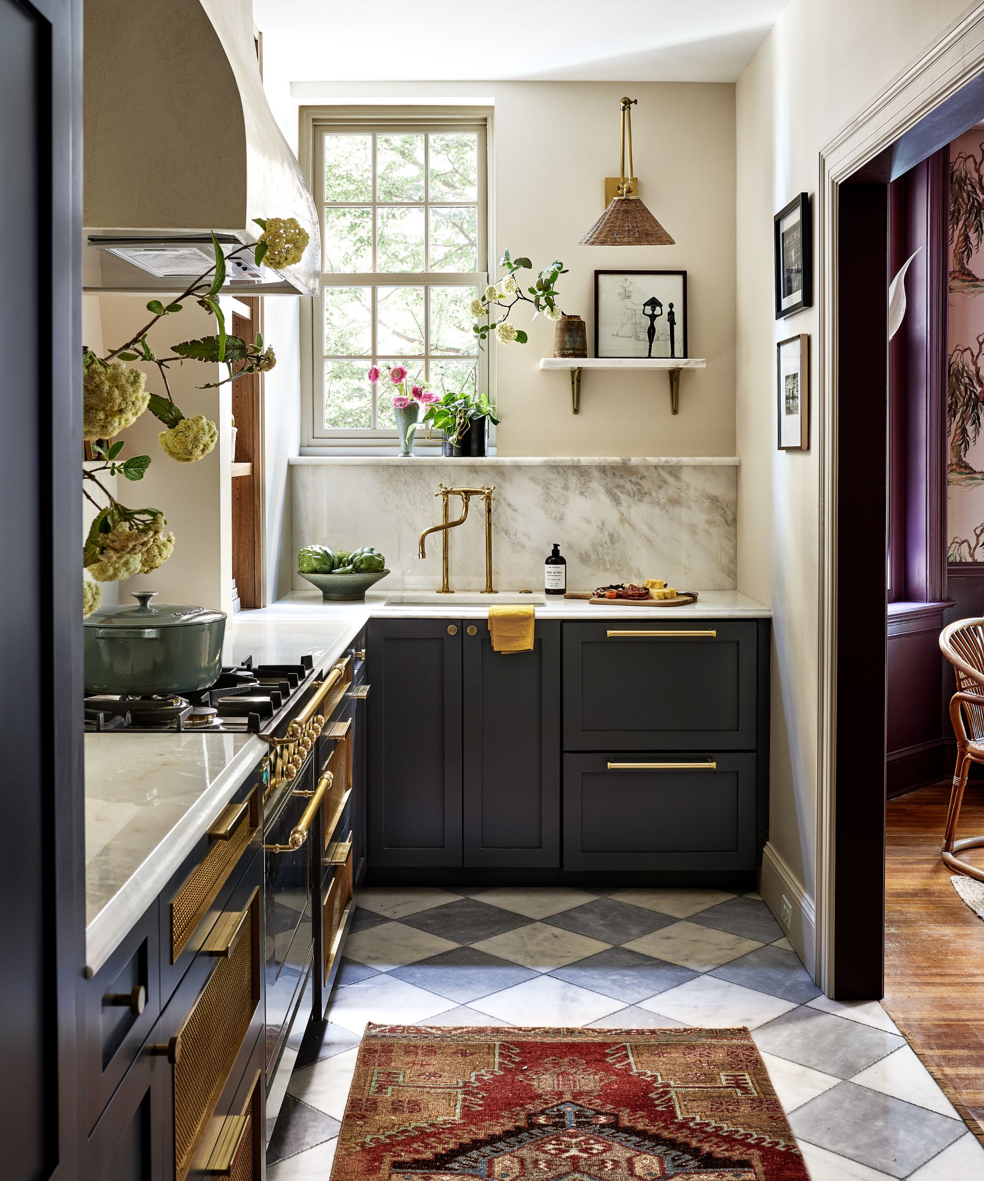 A small kitchen with dark blue cabinets, warm white walls, and a marble countersplash. A marble checkerboard floor is complemented by a red vintage-style runner, while vases of seasonal blooms are displayed throughout the room