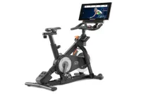 NordicTrack s22i Studio Cycle in this image is show with the rear of the bike on the left, and the front with a large integrated screen pointing to the right