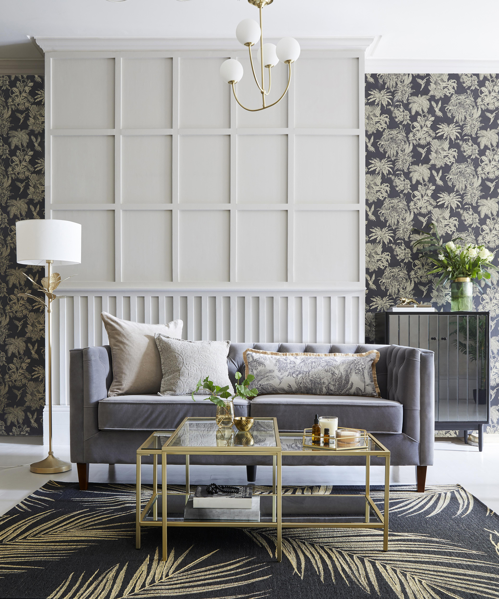 A grey and white living room with white wall paneling, charcoal grey and gold palm-print wallpaper, and grey velvet sofa