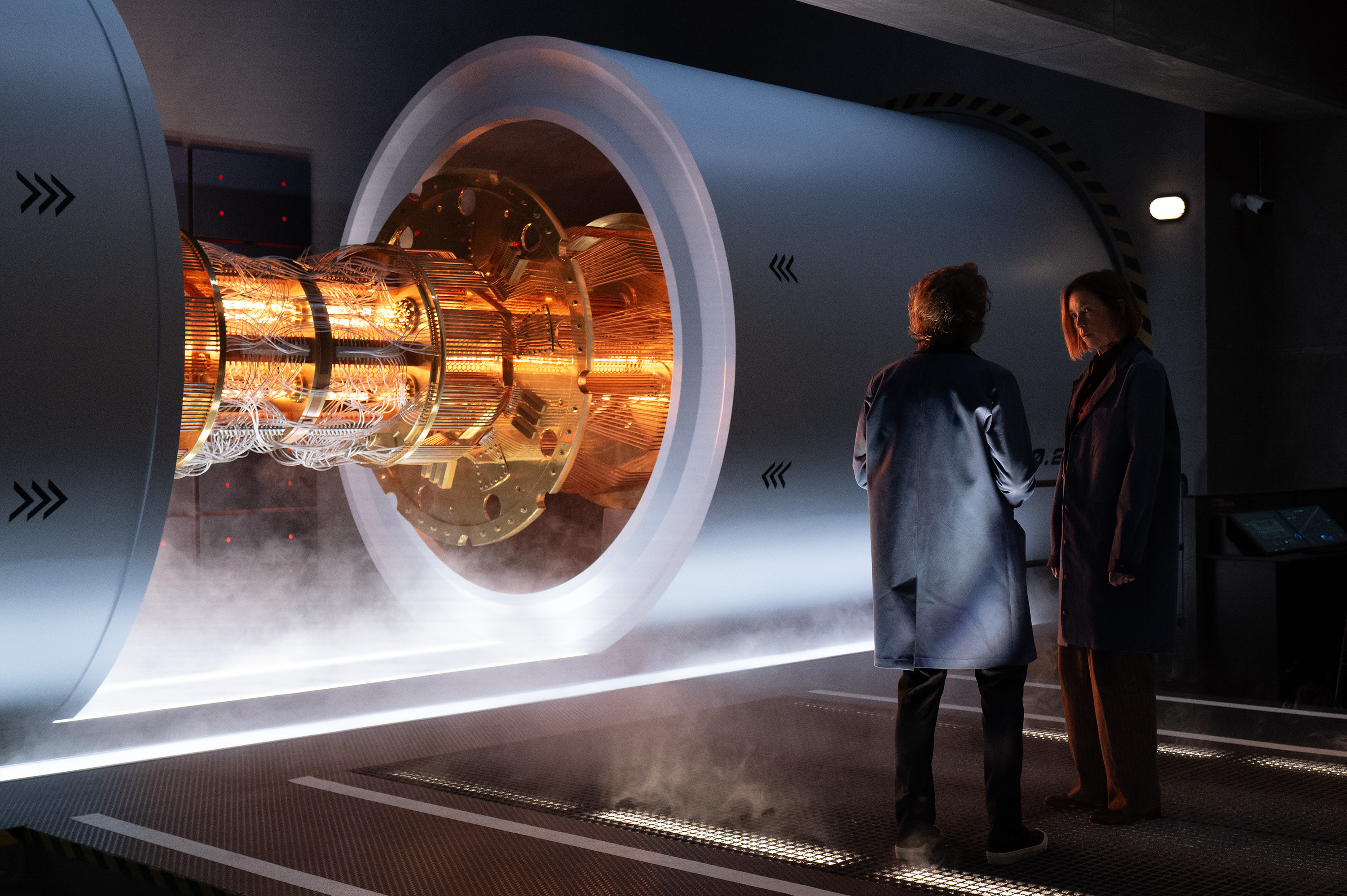 Two people (Simon Templeman as Dr. Chase and Julianne Nicholson as Sinatra) talk while standing to the right of a supercomputer, in a still from 'Paradise' season 2.