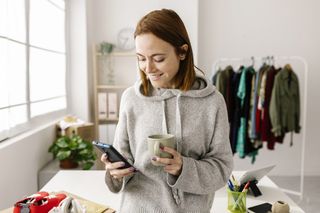 A smiling woman with a mug in one hand shops online from her smartphone