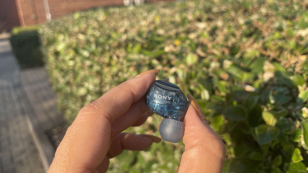A single Sony WF-C710N earbud being held with the eartip facing down in front of a hedge.