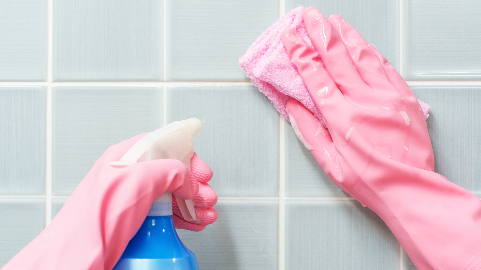 Rubber gloved hands holding a spray bottle and cloth against square tiles