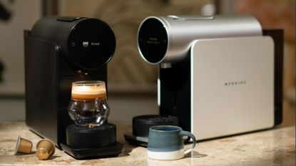 A black and a white Morning coffee maker on a kitchen counter, with one pouring a shot of espresso 