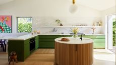 A bright, airy kitchen with a stone backsplash, kitchen drawers in green, a floating shelf, a wood-panelled round island, a wood stool, a door to the outside, and a white dining table with purple chairs by a colorful painting