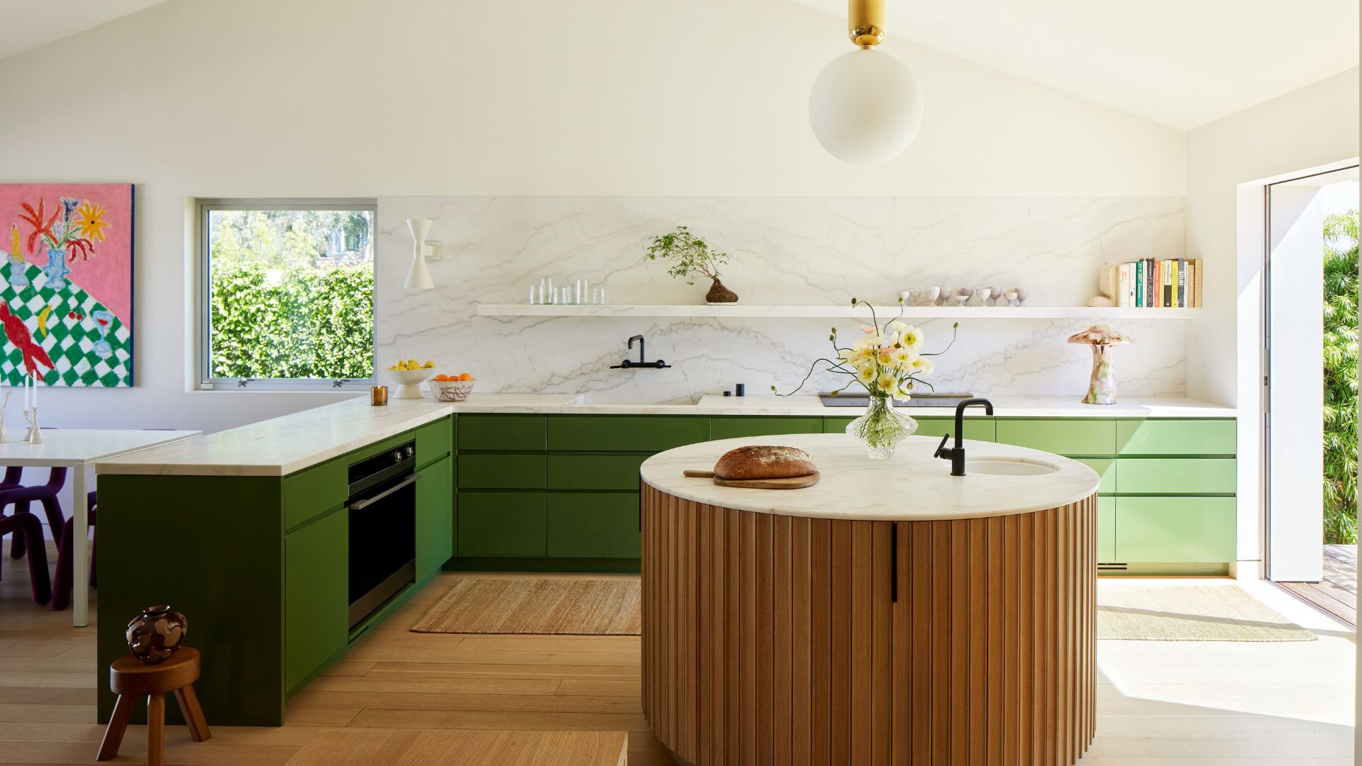 A bright, airy kitchen with a stone backsplash, kitchen drawers in green, a floating shelf, a wood-panelled round island, a wood stool, a door to the outside, and a white dining table with purple chairs by a colorful painting