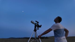 A man wearing a white and blue striped shirt stands next to a white telescope on a tripod as they both look up into a dark blue night sky where a bright dot is seen.
