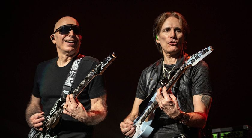 Joe Satriani wears wraparound shades as he jams onstage with old friend Steve Vai. Both are fretting a barre chord, both playing their signature Ibanez guitars.