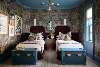 A bedroom with two single beds, patterned wallpaper, and blue storage trunks