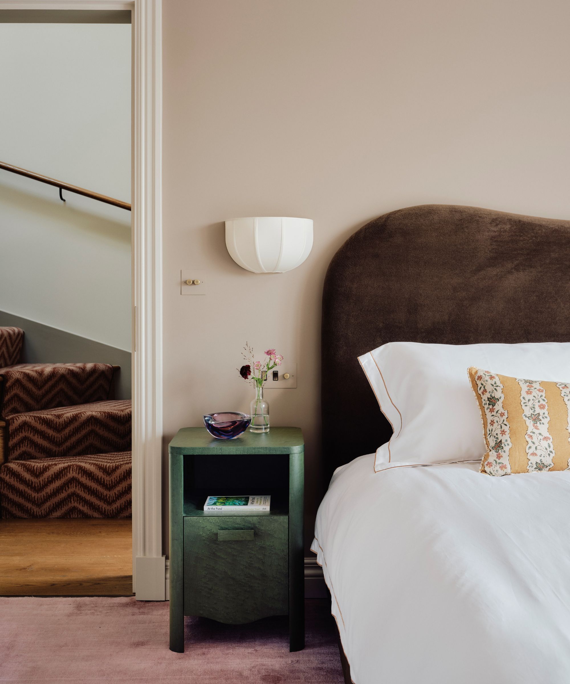 warm pink bedroom with a velvet chocolate brown headboard, white bed linen, a yellow striped floral cushion, green bedside table and lilac carpet