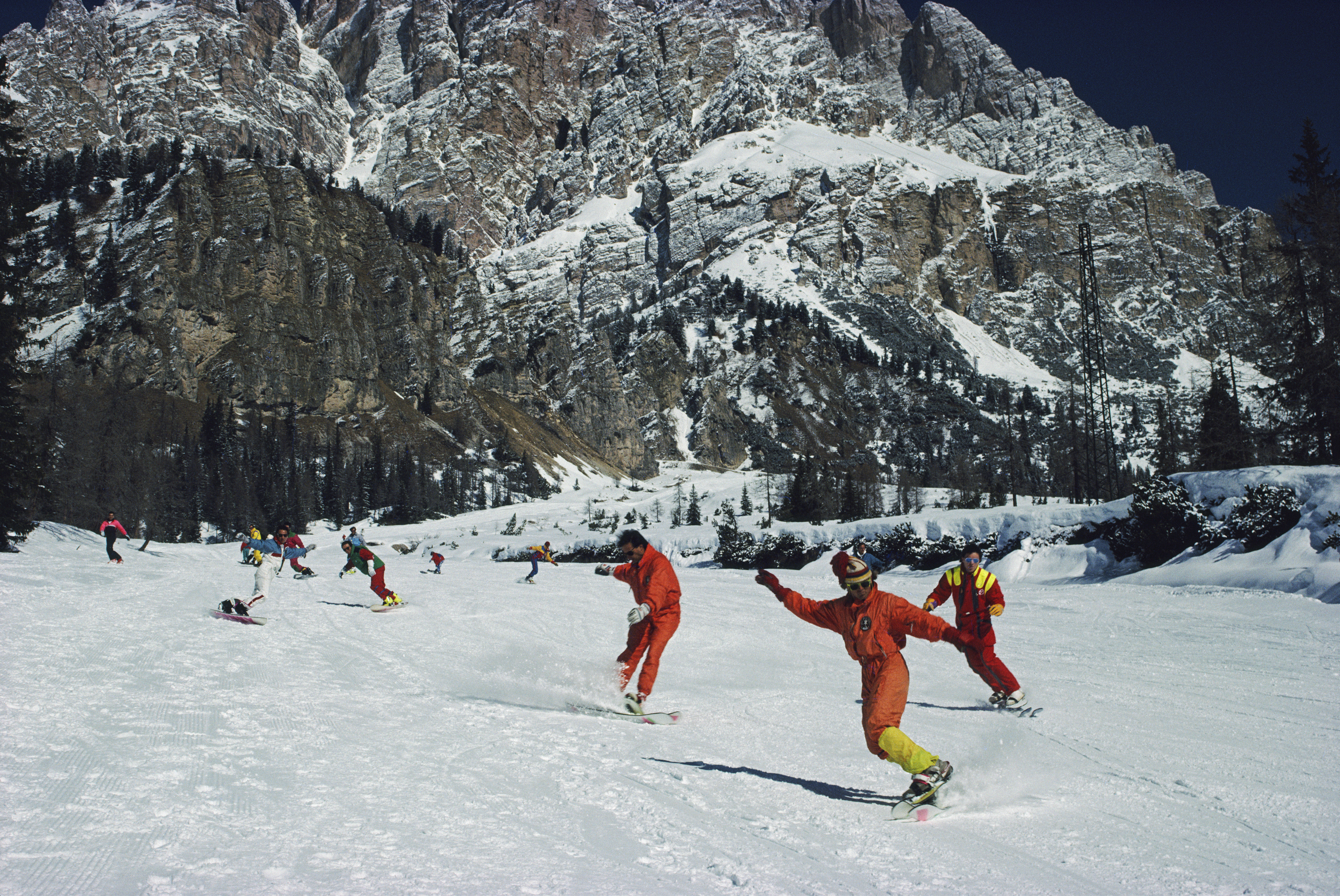 Snowboarders descending a wide alpine slope in colourful ski suits, carving through fresh snow beneath steep, rocky mountain cliffs.