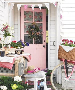 A white house and pink front door with a floral wreath and bunting above it, with a table in front of it with flowers, jam jars, and a basket on it, with a stool and a bike to the right hand side