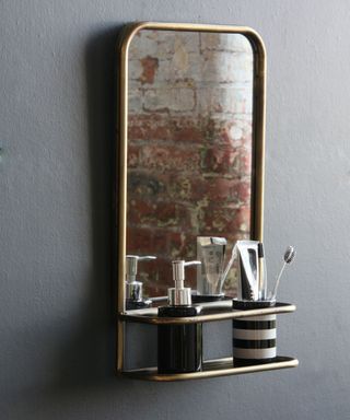 A dark gray bathroom wall with a gold metal shelf with a black soap dispenser and a black and white striped toothbrush pot, with the mirror reflecting a brick wall