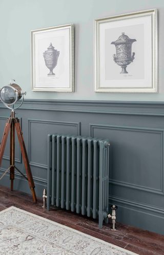 A dark blue painted radiator that matches wall paneling behind. A lighter blue painted on the top half of the wall, with two framed prints of urns hung up.