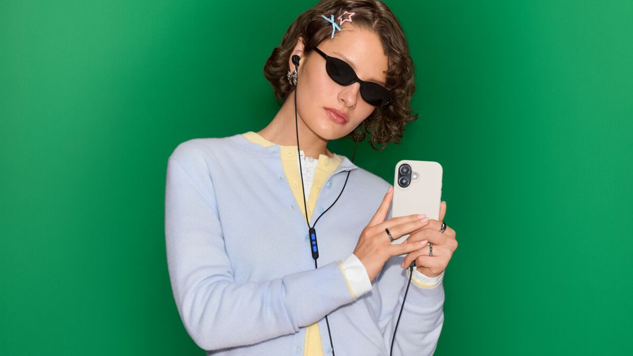 A young woman in a lilac cardigan and sunglasses holds her phone while wearing Bose Noise Cancelling Wired Earbuds