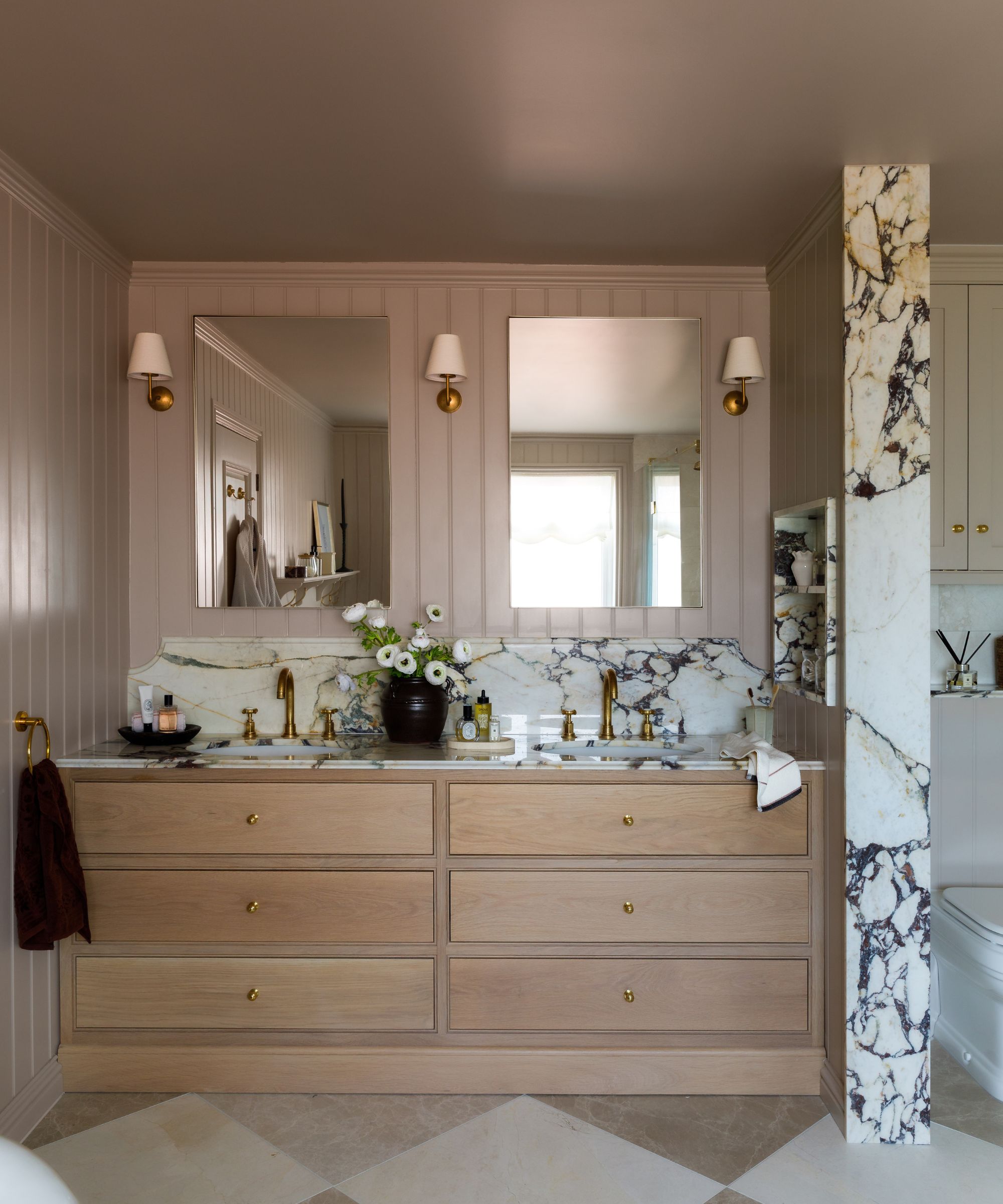 Bathroom with Elephant's Breath by Farrow &amp;amp; Ball painted walls, a double-sink vanity unit with marble countertop, wooden drawers built in underneath and two mirrors with sconces on either side.