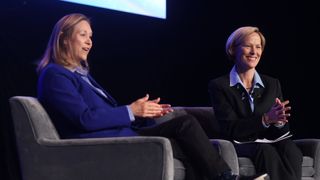 two women in blue suits sit on couches on a large stage