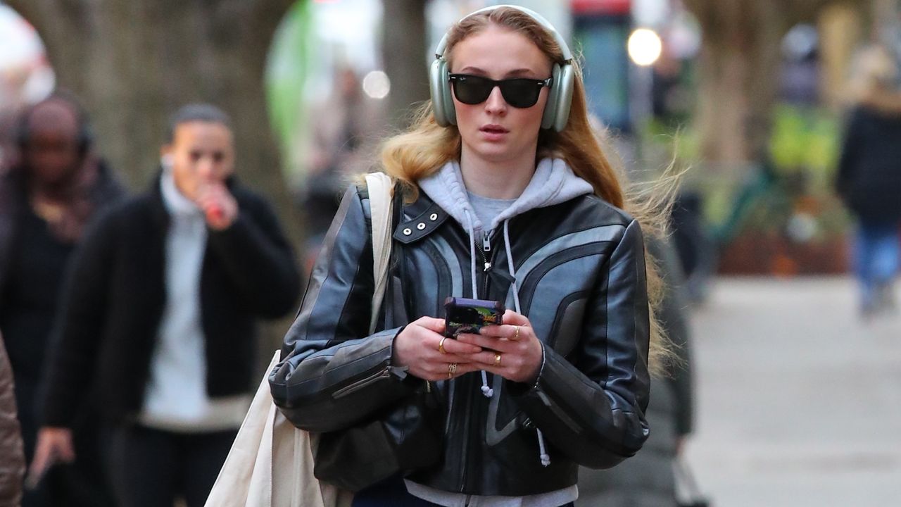 Sophie Turner in a leather jacket, blue pants, and a hoodie