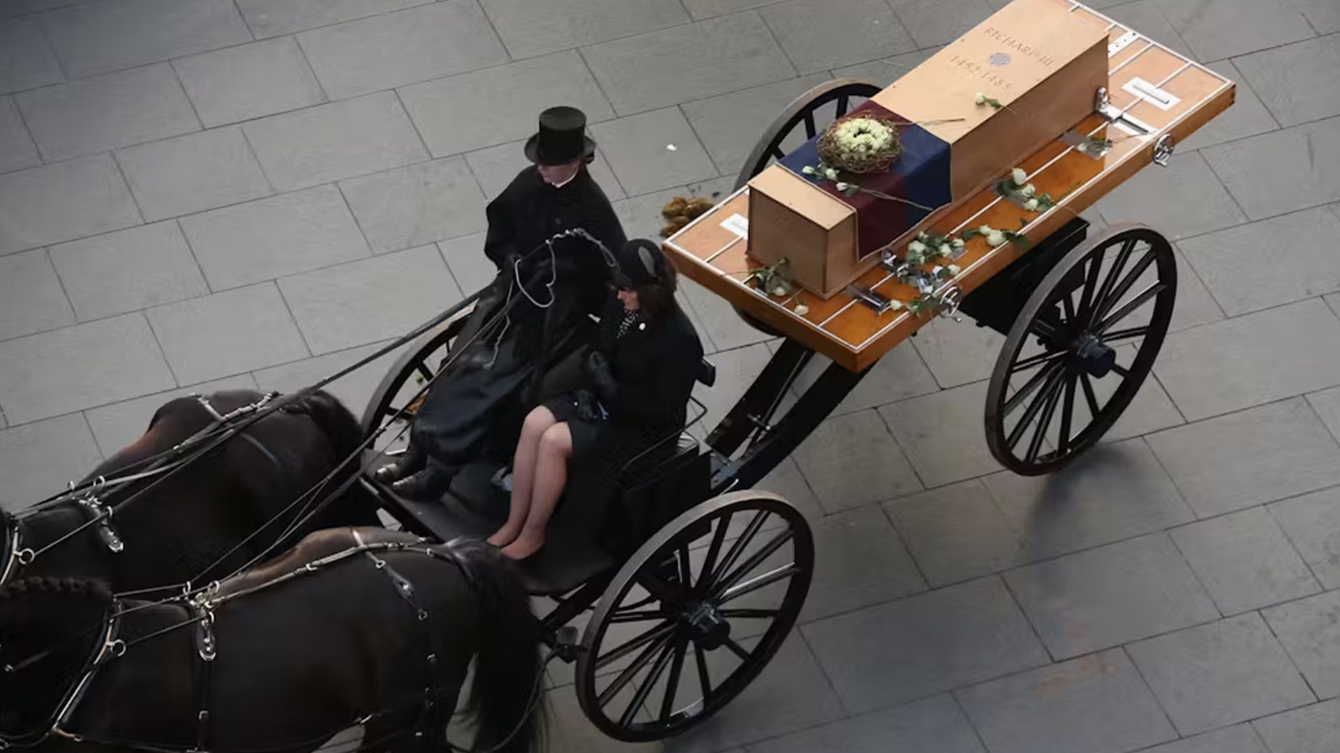 horse and carriage shown pulling a coffin on a flat platform; two people dressed in black are sitting in the open carriage