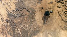 A satellite photo showing a black blob of ancient lava surrounding a volcano in the middle of the Sahara desert