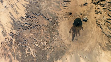 A satellite photo showing a black blob of ancient lava surrounding a volcano in the middle of the Sahara desert