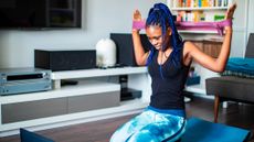 Woman kneels in front of laptop in living room. She holds a taut resistance band behind her head