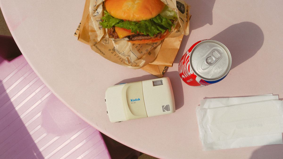 Photography of the Kodak EC35 film camera in white on a table beside a burger and a can of Coke