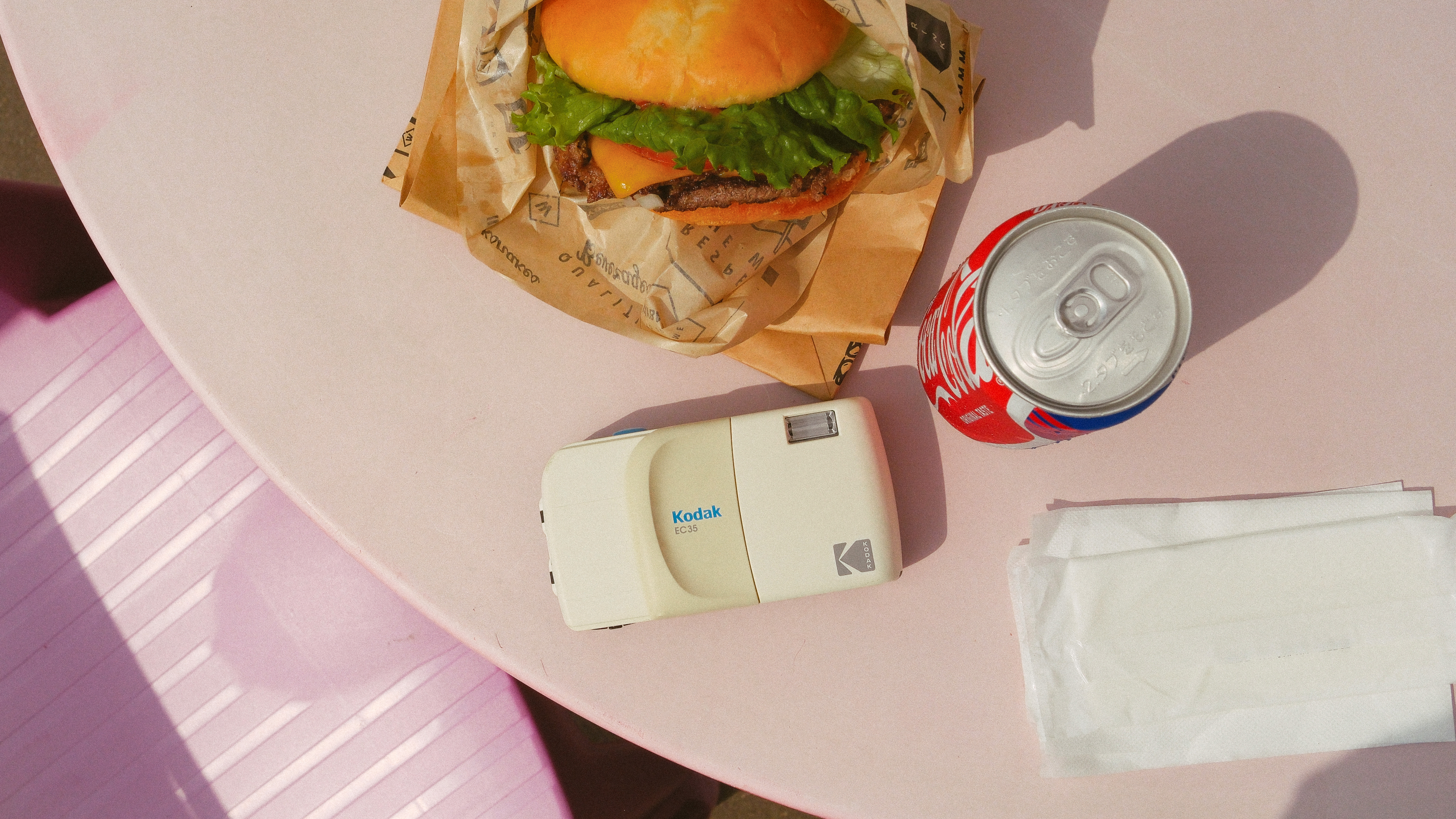 Photography of the Kodak EC35 film camera in white on a table beside a burger and a can of Coke