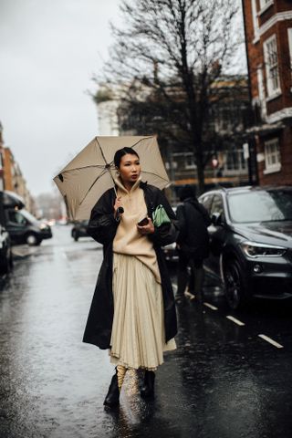 A woman wearing a knit sweater outfit of a tan zip-up sweater, black coat, pleated skirt, and boots