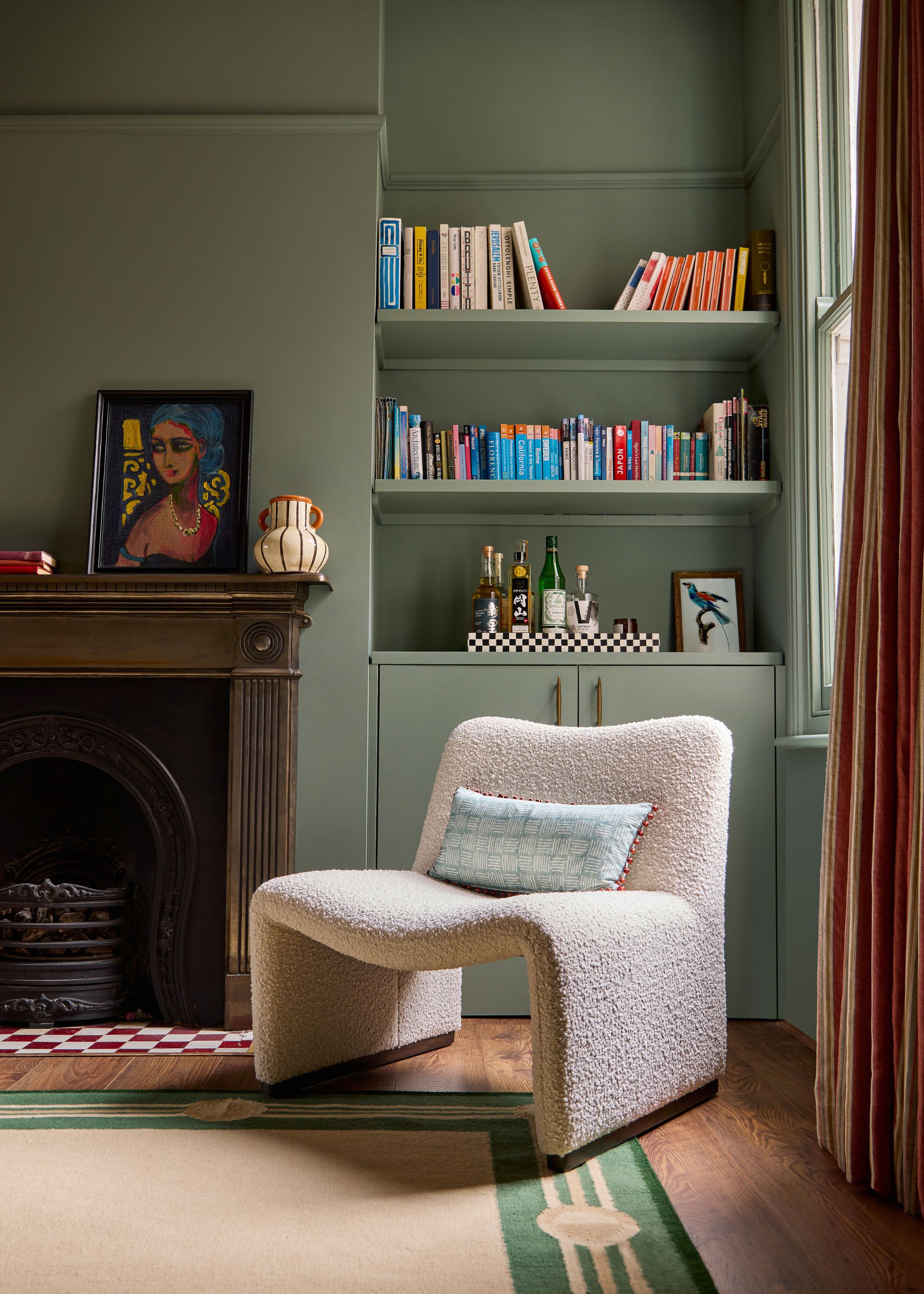 A living room in a period home with gray-green walls and a fireplace with a bronze mantel. Wooden flooring with a beige and green rug, a modern, cream boucle chair, and alcove shelving with books.